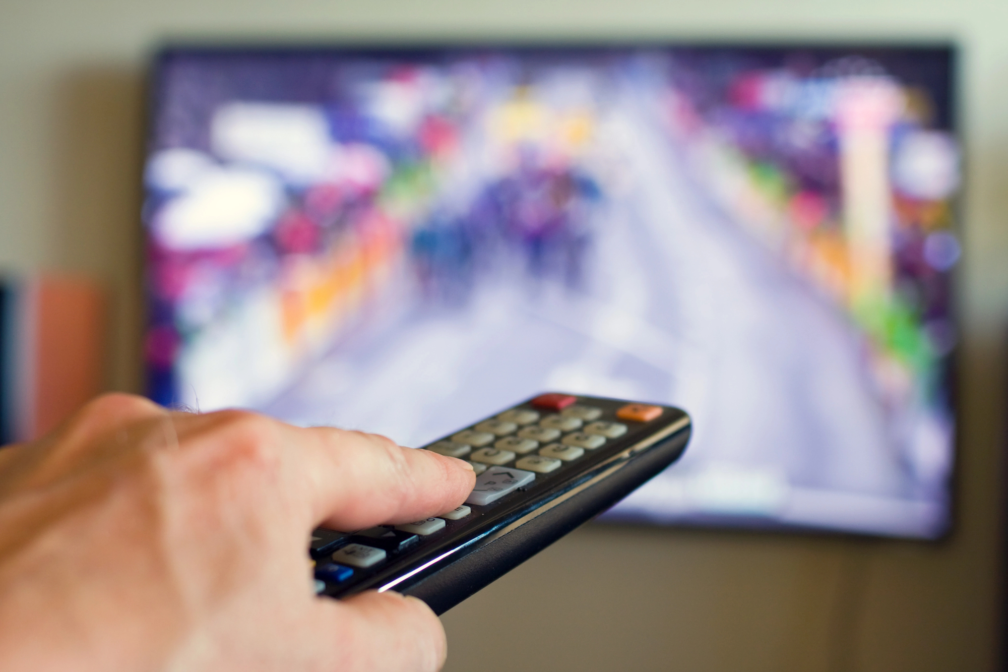 A hand holding a TV remote points towards a television displaying a blurred image of a colorful outdoor scene, possibly a race or parade, with people and banners on the screen.