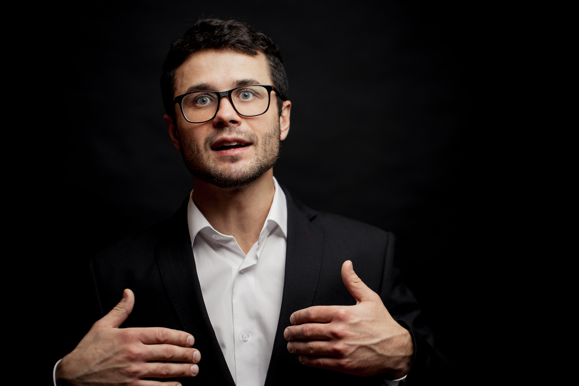 A man with glasses, dark hair, and a beard wears a black suit and a white shirt. He is gesturing with both hands against a dark background, appearing to be speaking or presenting.