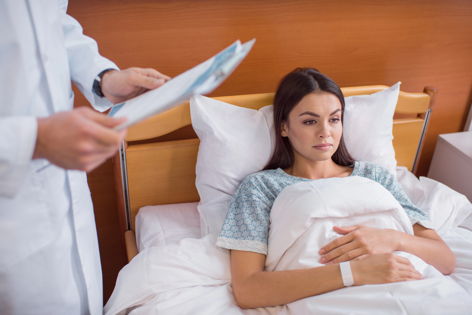 A woman lies in a hospital bed, looking pensive, covered with a white blanket. A doctor, holding a clipboard, stands nearby. The room has a wooden headboard and white bedding.