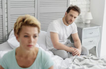 A couple sitting on a bed in a bedroom with a gray and white color scheme. The woman, with short blonde hair, looks away, appearing thoughtful. The man, with short brown hair, sits behind her, looking concerned. A bedside table with a lamp is visible.