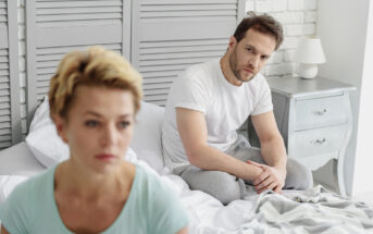 A couple sitting on a bed in a bedroom with a gray and white color scheme. The woman, with short blonde hair, looks away, appearing thoughtful. The man, with short brown hair, sits behind her, looking concerned. A bedside table with a lamp is visible.