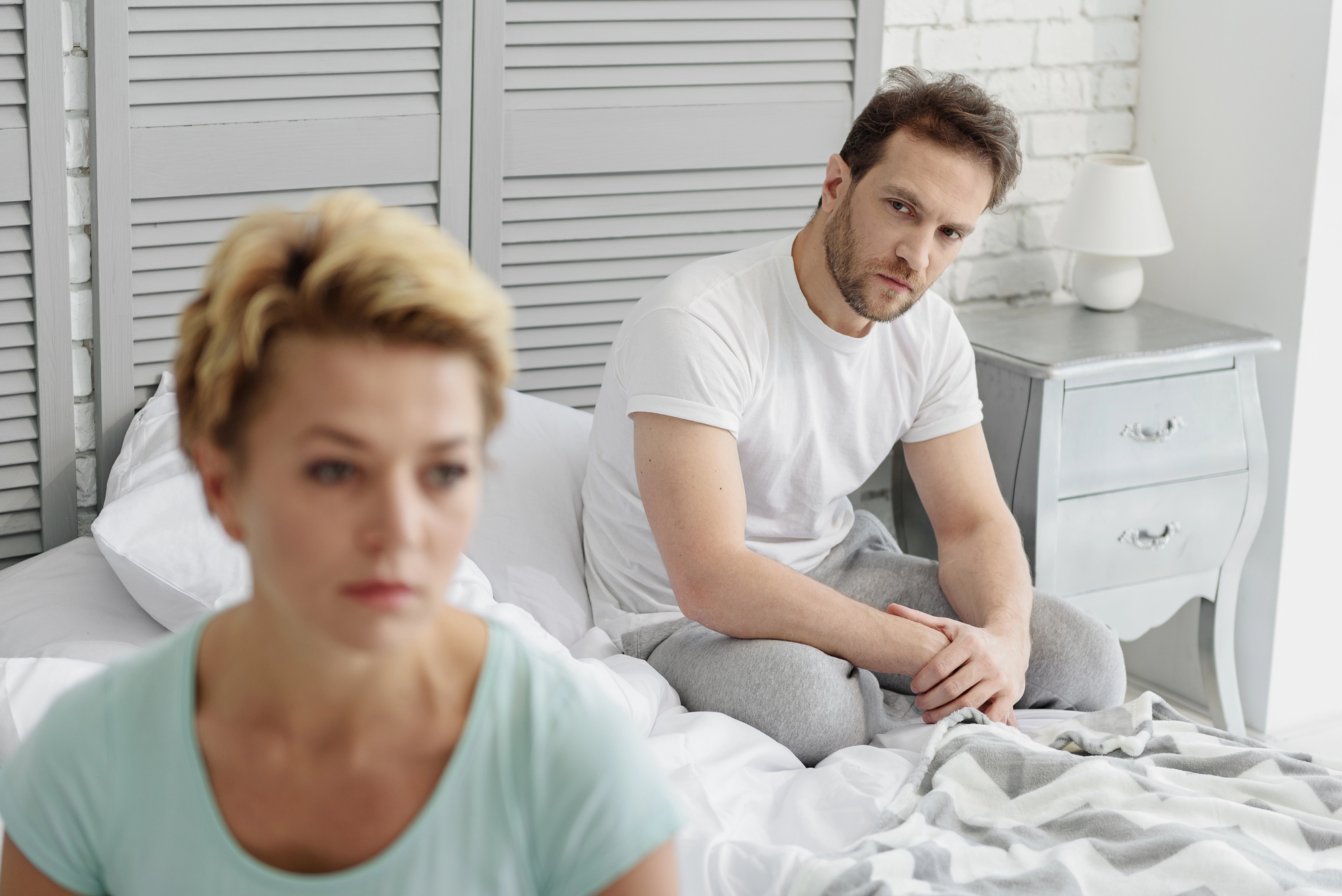 A couple sitting on a bed in a bedroom with a gray and white color scheme. The woman, with short blonde hair, looks away, appearing thoughtful. The man, with short brown hair, sits behind her, looking concerned. A bedside table with a lamp is visible.