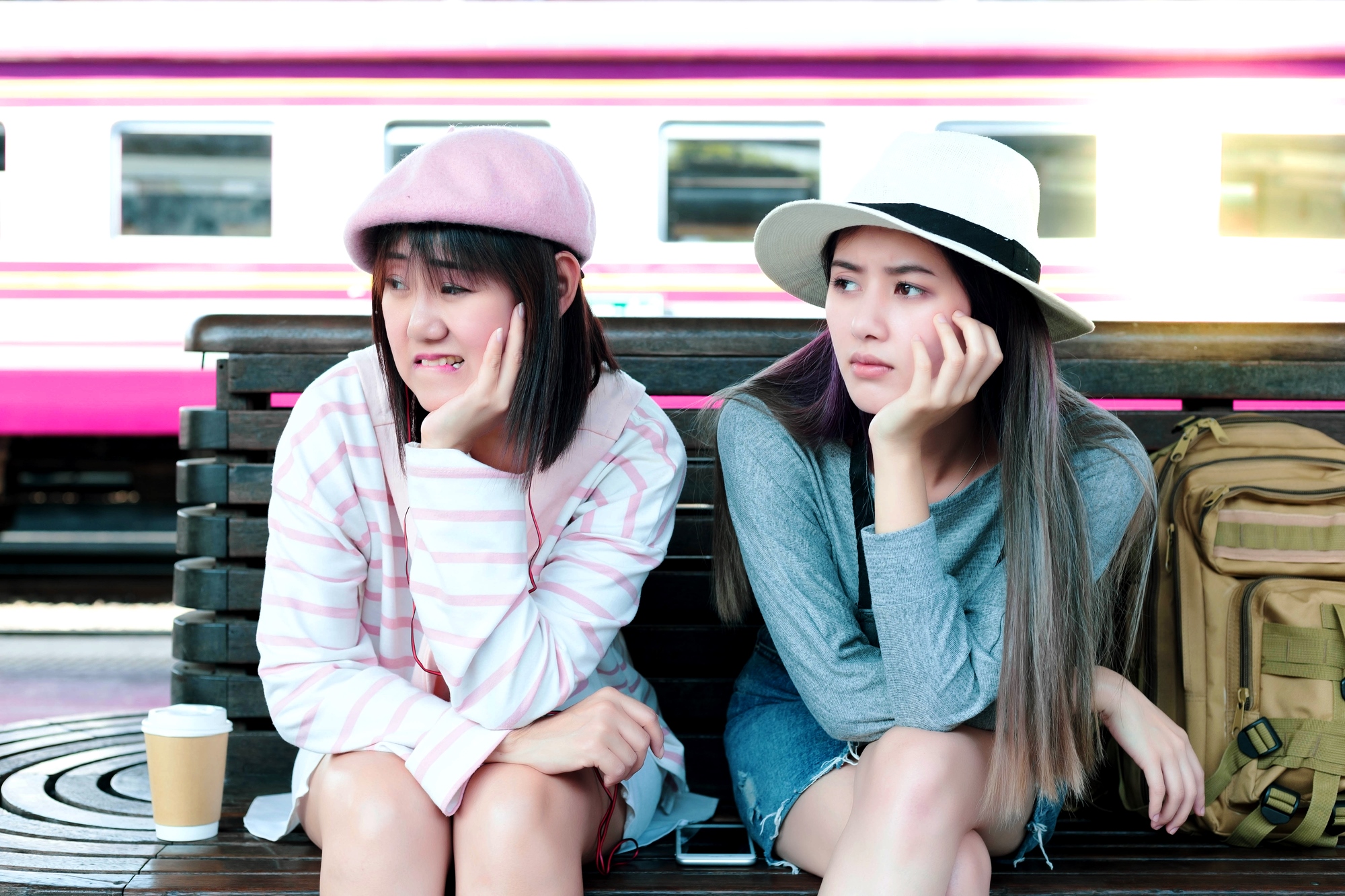 Two people sitting on a bench at a train station, looking bored. One wears a pink hat and striped shirt, the other a white hat and gray sweater. A backpack and coffee cup are beside them, and a moving train is blurred in the background.