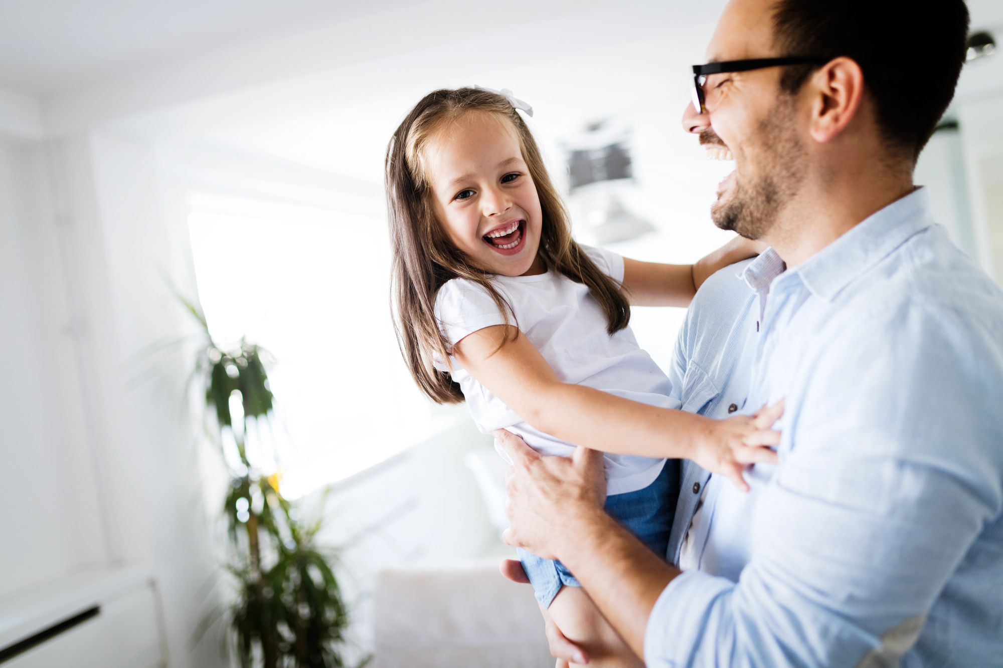 A man wearing glasses joyfully holds a smiling young girl in his arms in a bright living room. Both appear happy and relaxed. A potted plant is visible in the background, adding a touch of greenery to the scene.