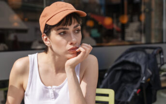 A person with short hair and an orange cap sits outside, resting their chin on their hand. They are wearing a white tank top. A stroller is visible in the background.