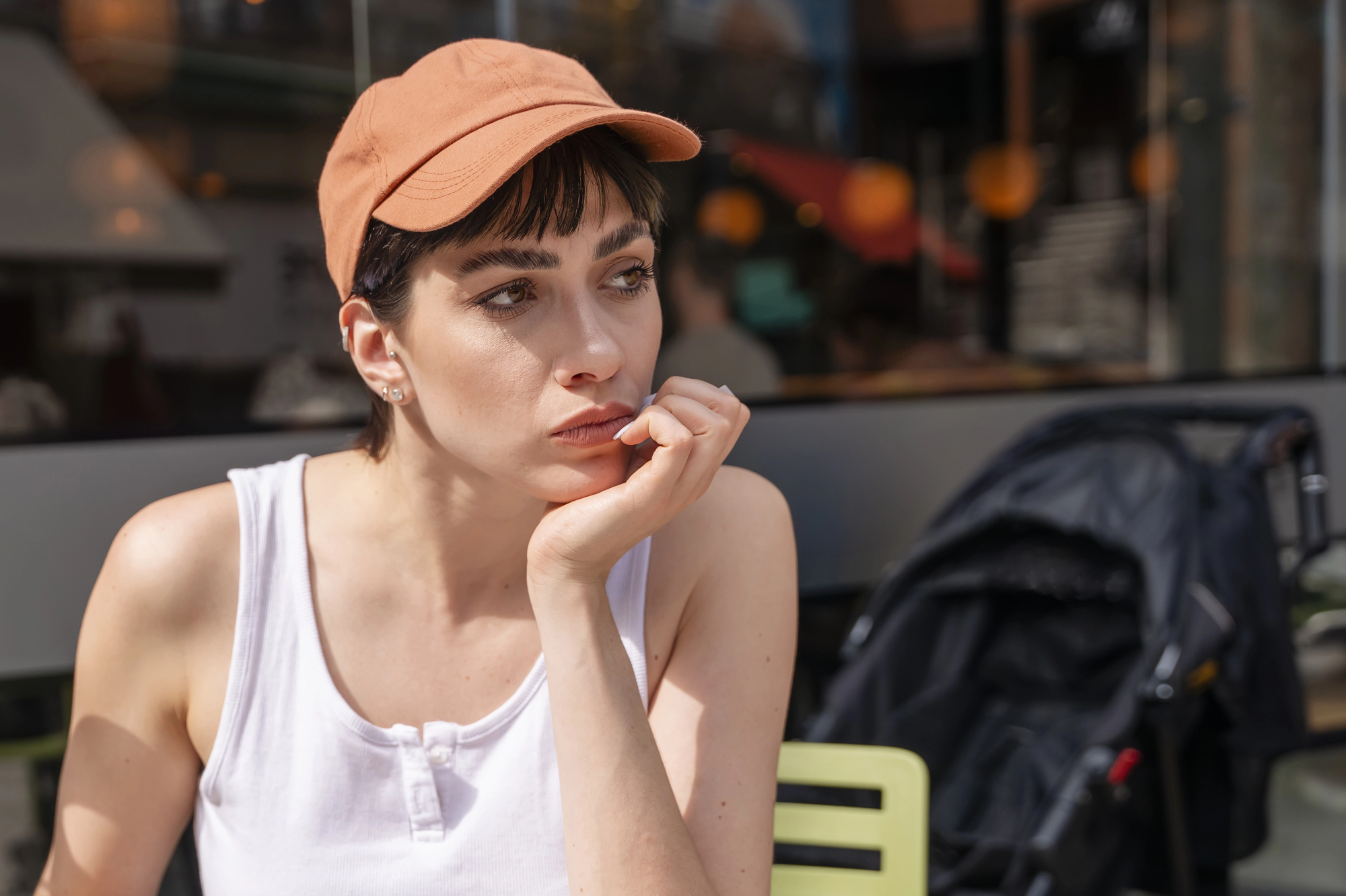 A person with short hair and an orange cap sits outside, resting their chin on their hand. They are wearing a white tank top. A stroller is visible in the background.