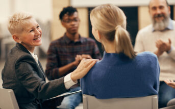 A woman with short white hair smiles and touches the shoulder of another woman wearing a blue sweater. They are seated in a circle with two other people, suggesting a supportive group meeting.