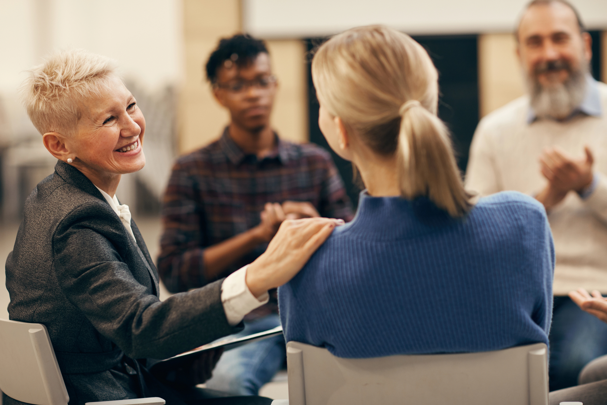 A woman with short white hair smiles and touches the shoulder of another woman wearing a blue sweater. They are seated in a circle with two other people, suggesting a supportive group meeting.