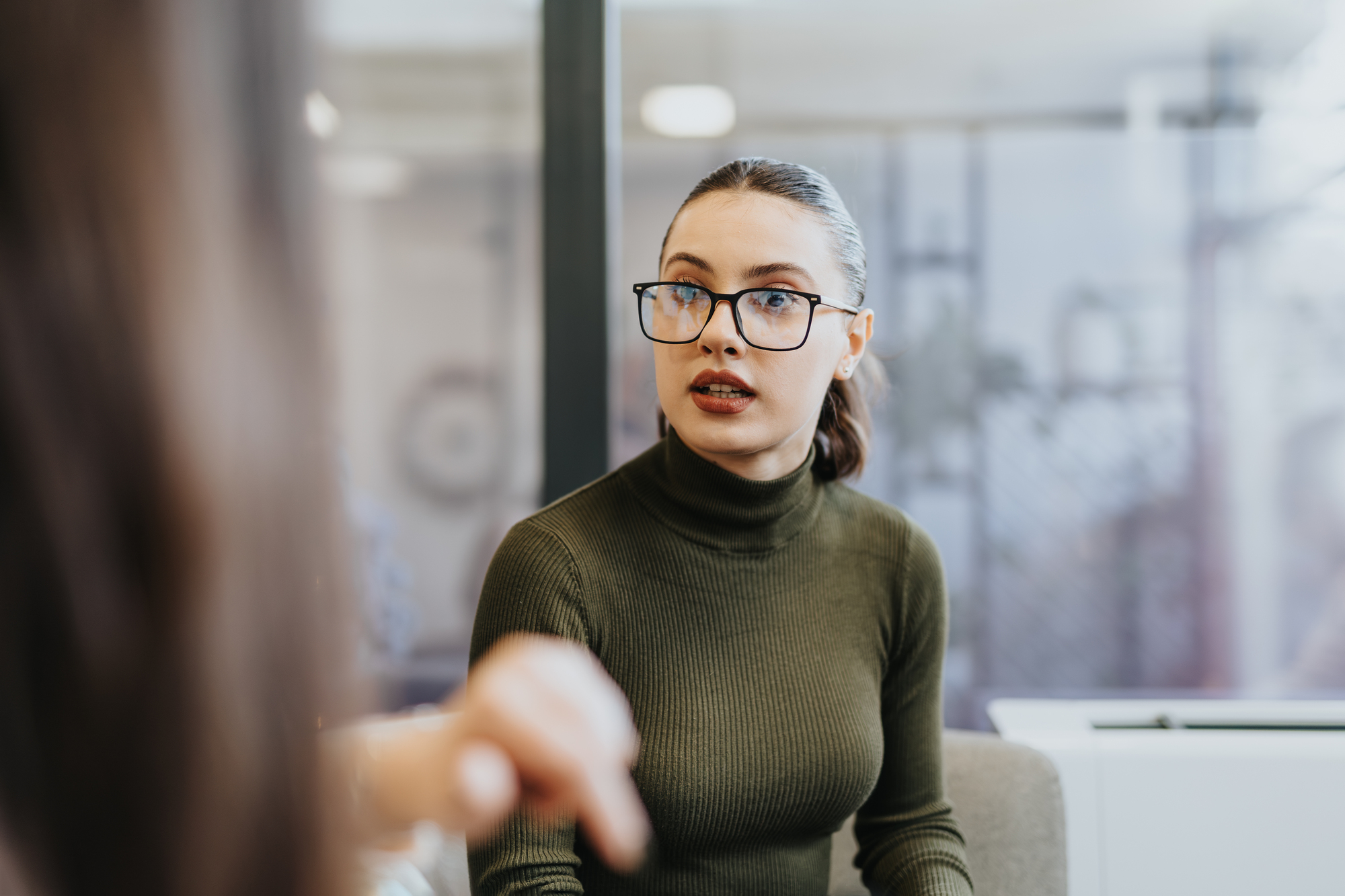 A woman with glasses and a ponytail sits indoors, wearing a green turtleneck. She appears engaged in conversation, with a blurred hand gesturing in the foreground. The setting seems to be an office or casual meeting space.