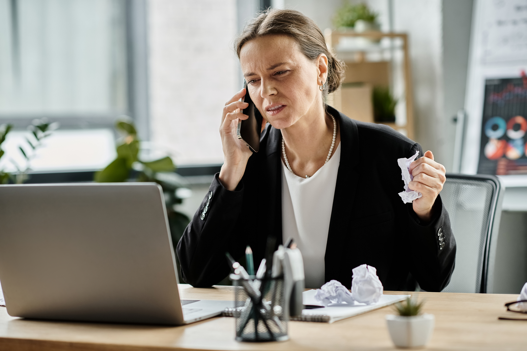 A person in a suit is sitting at a desk, holding a phone to their ear and a crumpled piece of paper in one hand. They are looking at a laptop screen with a concerned expression. The desk has pens and more crumpled papers on it.