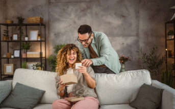 A woman with curly hair sits on a couch, smiling as she holds an open gift box. A man with glasses stands behind, looking over her shoulder with a joyful expression. The room has a cozy, modern decor with plants and shelves in the background.