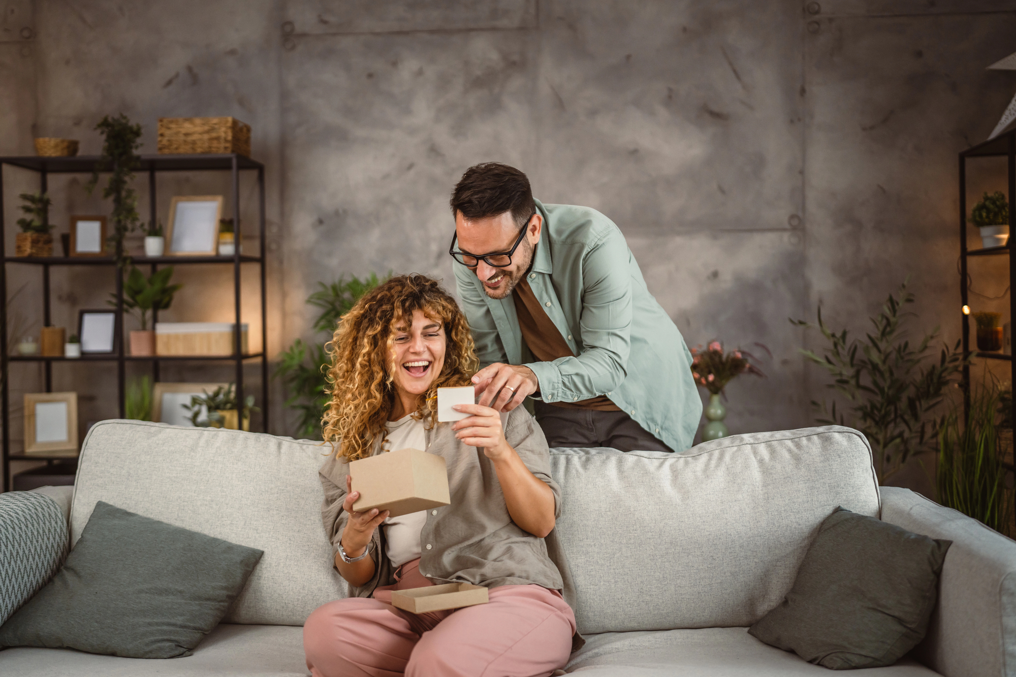 A woman with curly hair sits on a couch, smiling as she holds an open gift box. A man with glasses stands behind, looking over her shoulder with a joyful expression. The room has a cozy, modern decor with plants and shelves in the background.