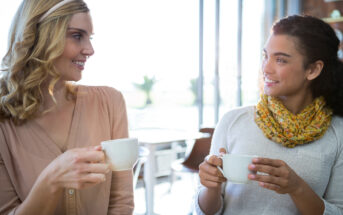 Two women are sitting in a bright, modern cafe, smiling and holding cups of coffee. One wears a beige blouse and the other a white sweater with a yellow scarf. They appear engaged in a friendly conversation.