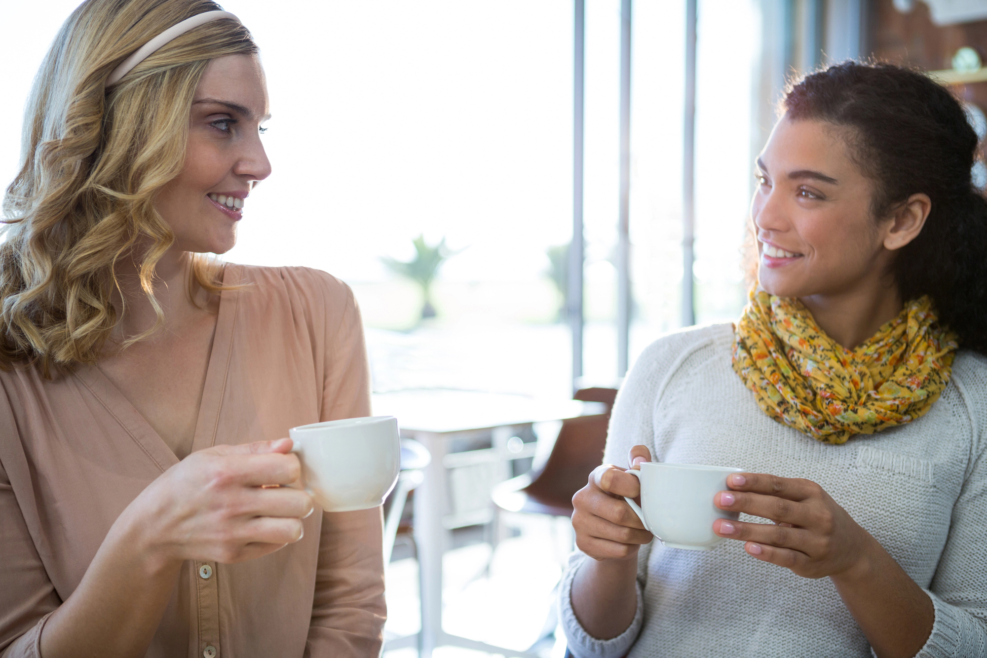 Two women are sitting in a bright, modern cafe, smiling and holding cups of coffee. One wears a beige blouse and the other a white sweater with a yellow scarf. They appear engaged in a friendly conversation.