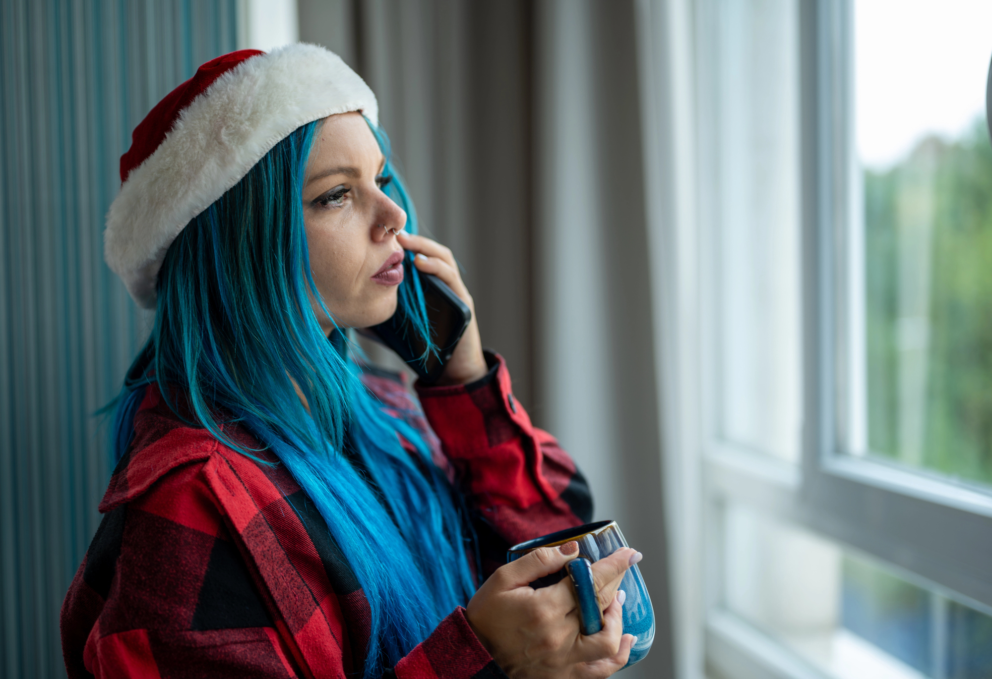 A woman with long blue hair wearing a Santa hat and red plaid shirt is talking on the phone while holding a mug. She is looking out of a window, with natural light illuminating her face.