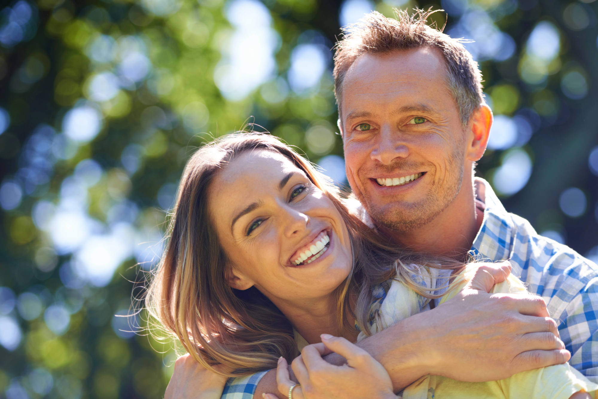 A smiling man and woman embraced in a park with dappled sunlight filtering through the trees. Both are casually dressed, and they appear happy and relaxed, surrounded by a lush, green background.