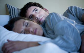 A man lies in bed, eyes open and appearing deep in thought, while a woman sleeps peacefully beside him. They are both covered with a striped duvet. The room has a soft, calming light.