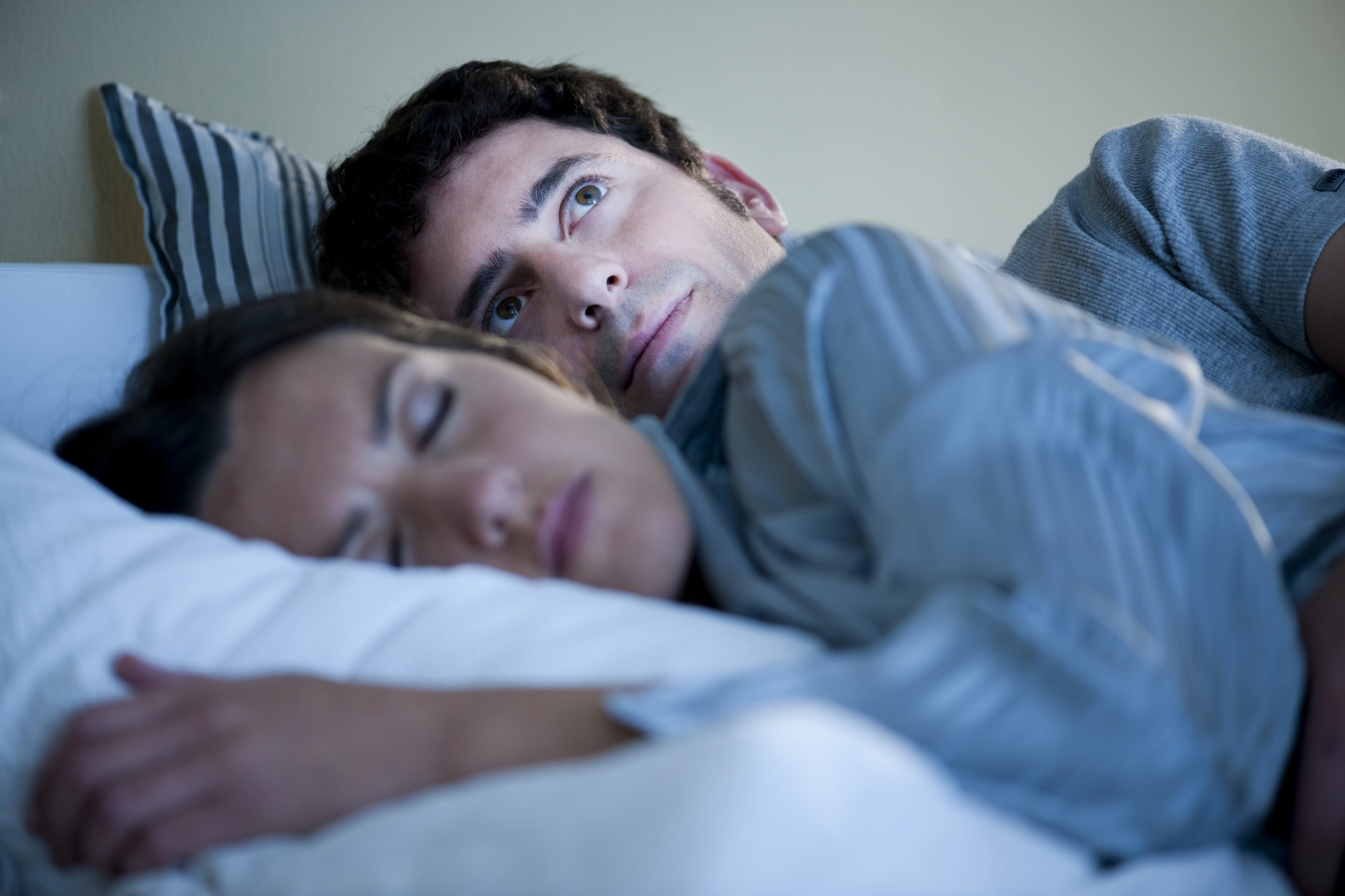 A man lies in bed, eyes open and appearing deep in thought, while a woman sleeps peacefully beside him. They are both covered with a striped duvet. The room has a soft, calming light.