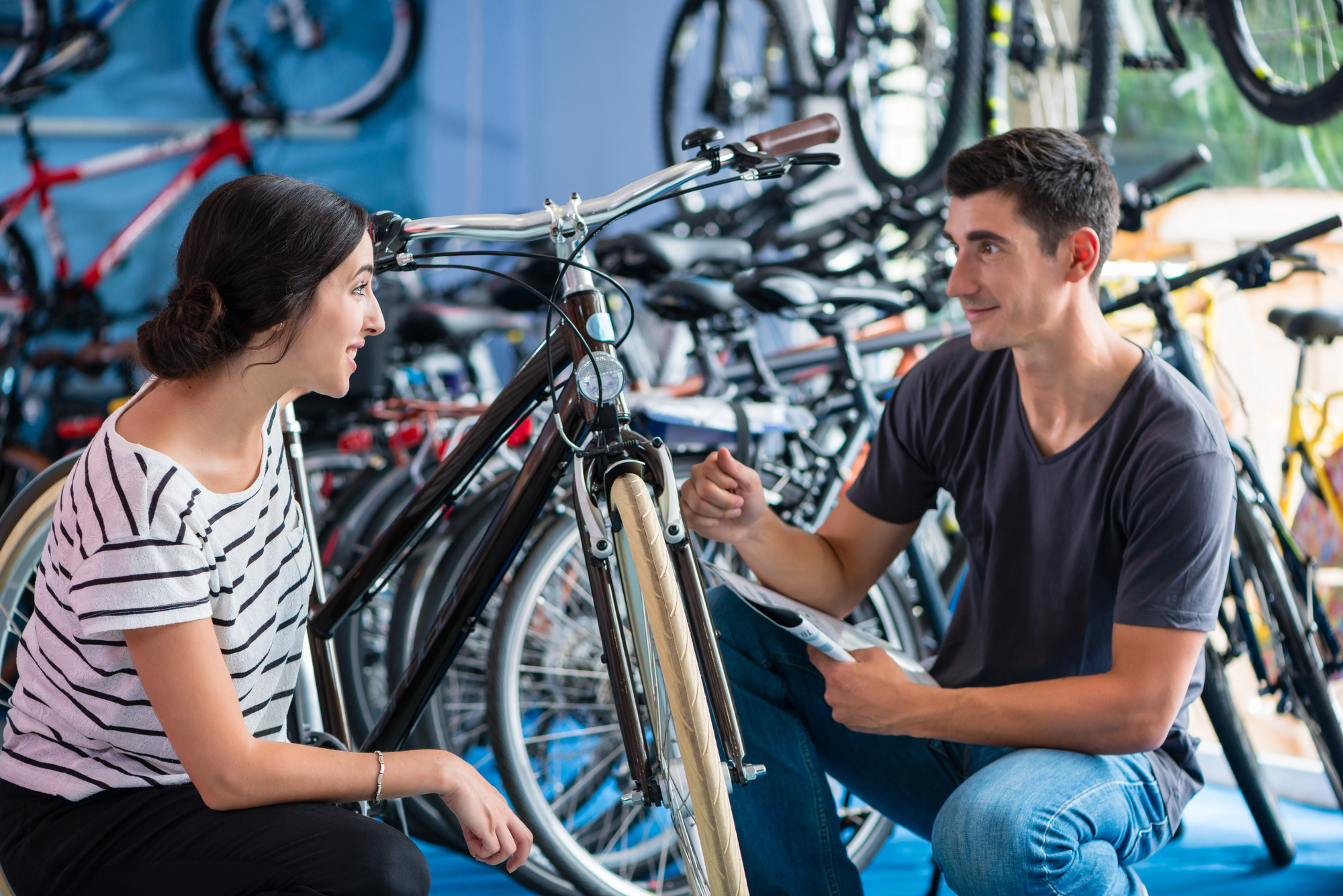 A woman and a man are in a bike shop. The man is kneeling and holding a brochure, discussing a bicycle with the woman, who is crouching beside him. The shop is filled with various bicycles in the background.