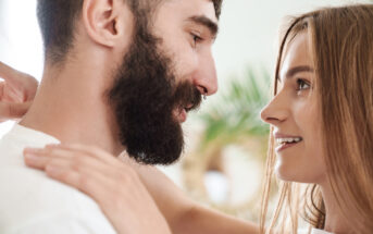 A man with a beard and a woman with long hair are smiling at each other closely. The background is softly blurred, with a hint of greenery. They appear to be in a joyful or intimate moment.