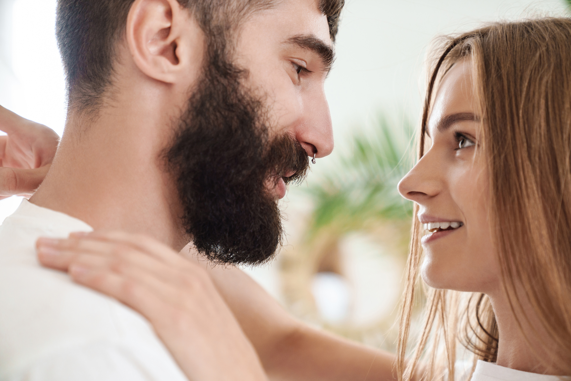 A man with a beard and a woman with long hair are smiling at each other closely. The background is softly blurred, with a hint of greenery. They appear to be in a joyful or intimate moment.