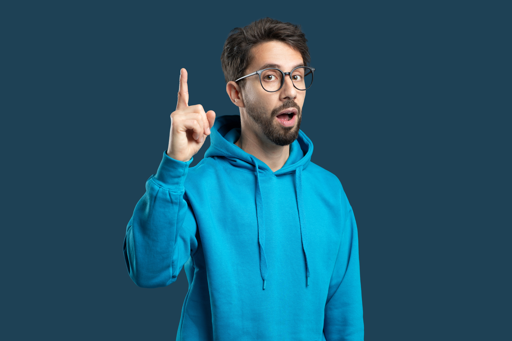 A man with glasses and a beard, wearing a blue hoodie, stands against a plain blue background. He is raising his index finger and has an expression of surprise or realization.