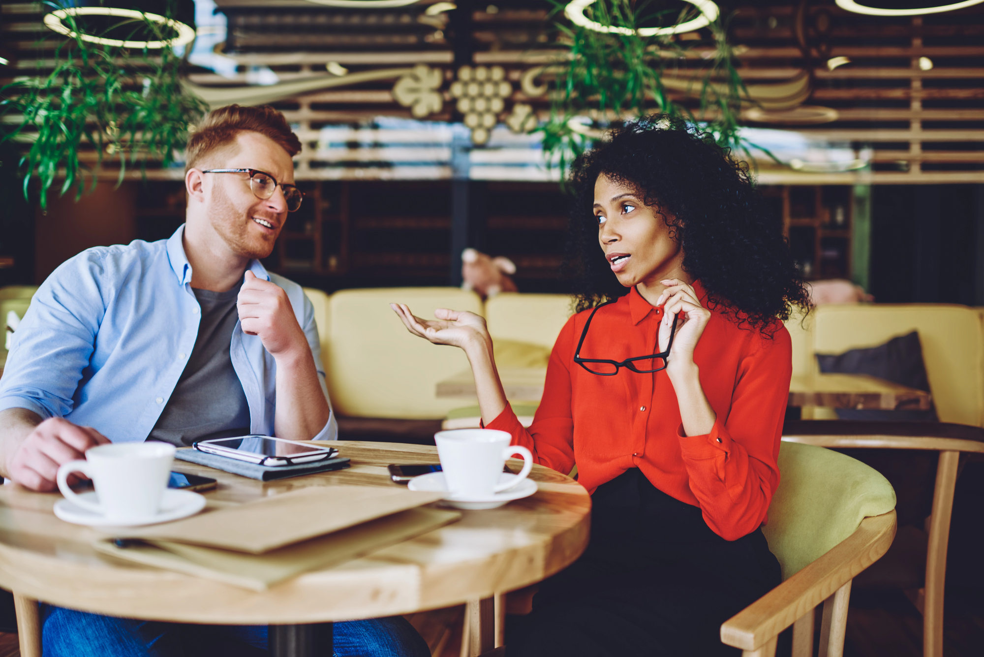 Two people sit at a café table with coffee cups and tablets. One person, in a light blue shirt, listens intently, while the other, in a red blouse, gestures expressively. Green plants hang from the ceiling, adding a touch of nature to the modern setting.