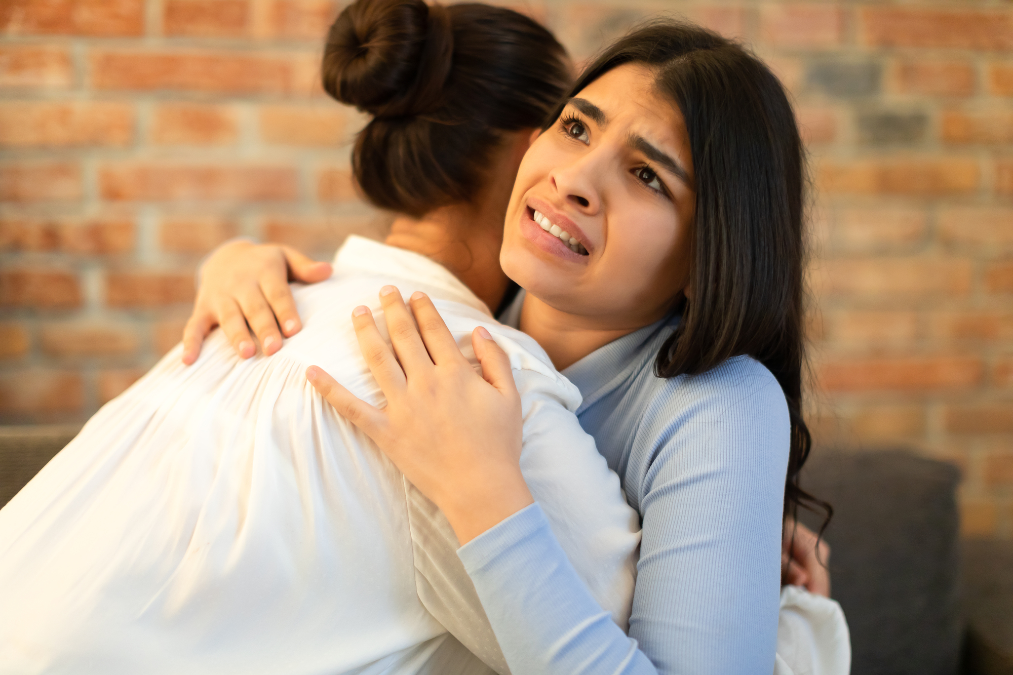 Two women hugging indoors. One facing the camera looks upset while the other has her back to the camera. The background features a brick wall, creating a cozy atmosphere.