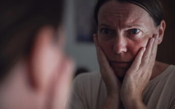 A person looks intently at their reflection in a mirror, with their hands on their cheeks, appearing deep in thought or concern. The soft lighting and close-up perspective emphasize their expression.