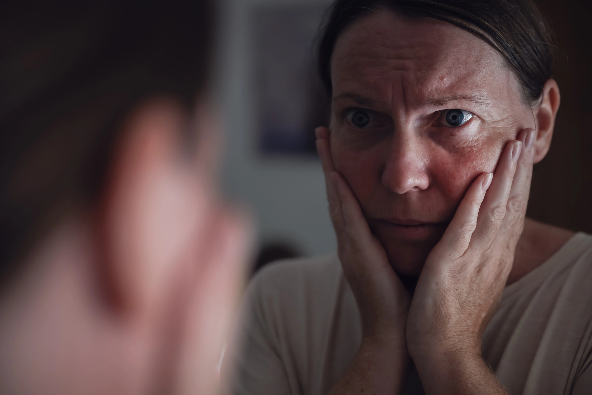 A person looks intently at their reflection in a mirror, with their hands on their cheeks, appearing deep in thought or concern. The soft lighting and close-up perspective emphasize their expression.