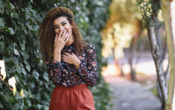 A woman with curly hair stands near a lush ivy-covered wall. She is smiling and covering her mouth with one hand, wearing a floral blouse and rust-colored pants. Sunlight filters through trees in the background, creating a warm, pleasant atmosphere.