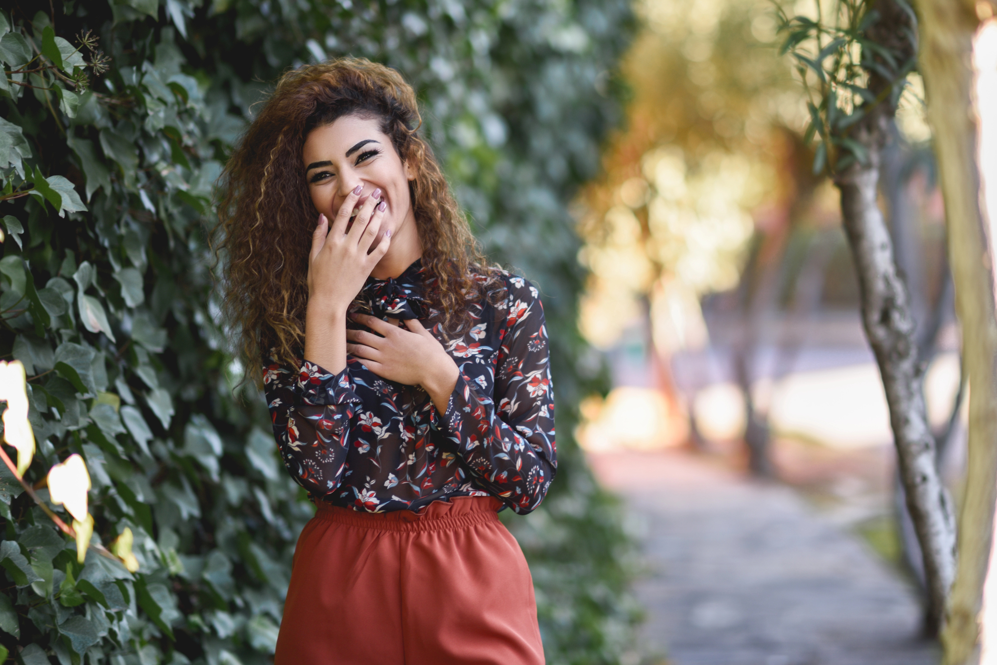 A woman with curly hair stands near a lush ivy-covered wall. She is smiling and covering her mouth with one hand, wearing a floral blouse and rust-colored pants. Sunlight filters through trees in the background, creating a warm, pleasant atmosphere.