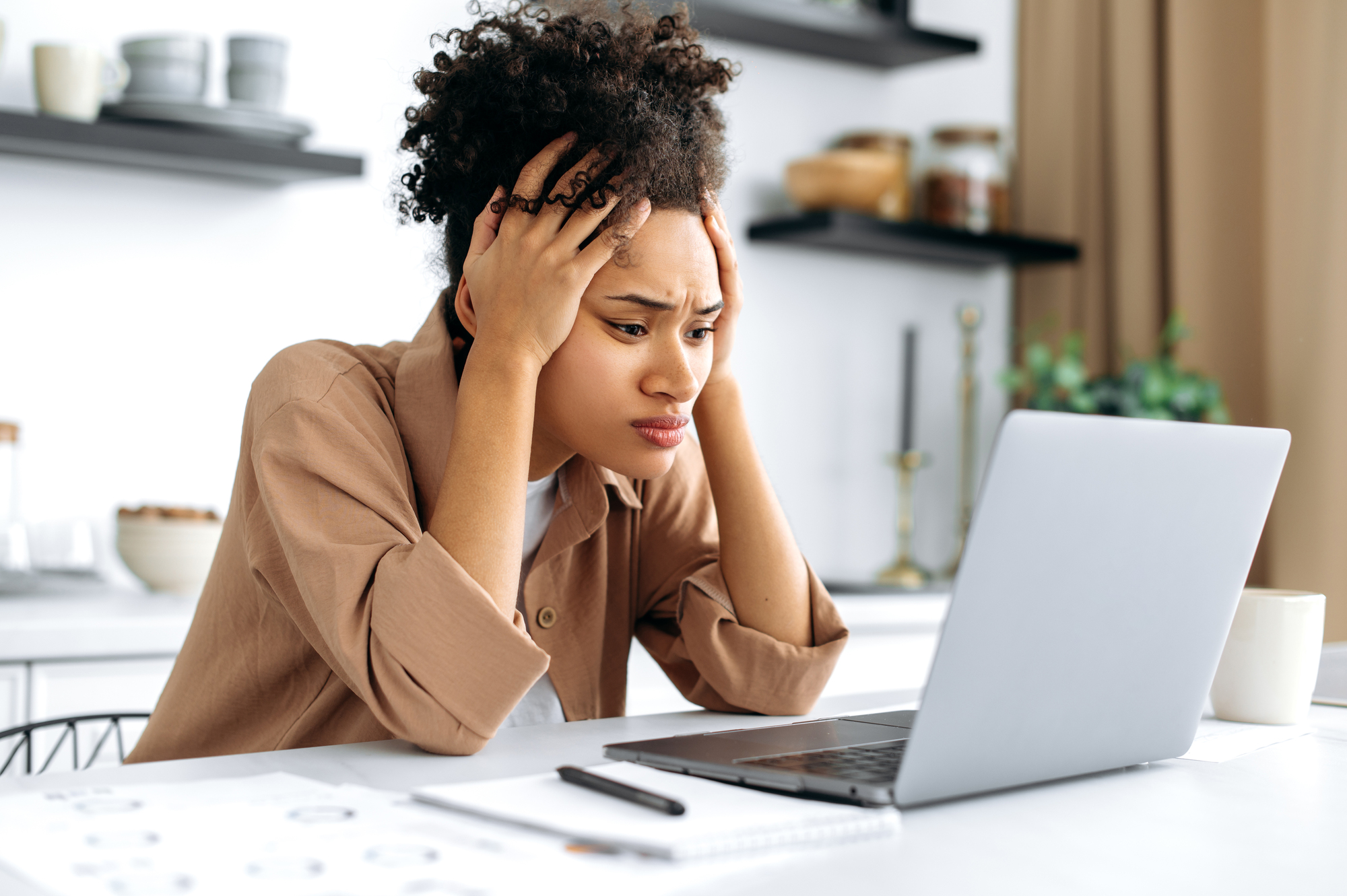 A woman with curly hair sits at a kitchen table, looking stressed while working on a laptop. She holds her head with both hands, and papers, a notebook, and a pen are scattered around her. Shelves with jars are visible in the background.