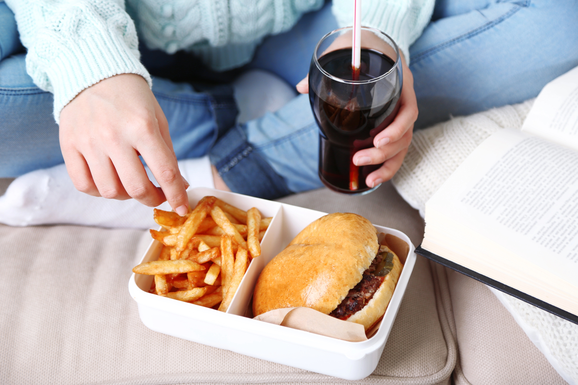 A person sitting cross-legged, holding a glass of cola with a straw. In front of them is an open book and a lunch container with a burger and fries. They are reaching for a fry with one hand. They are wearing jeans and a light sweater.