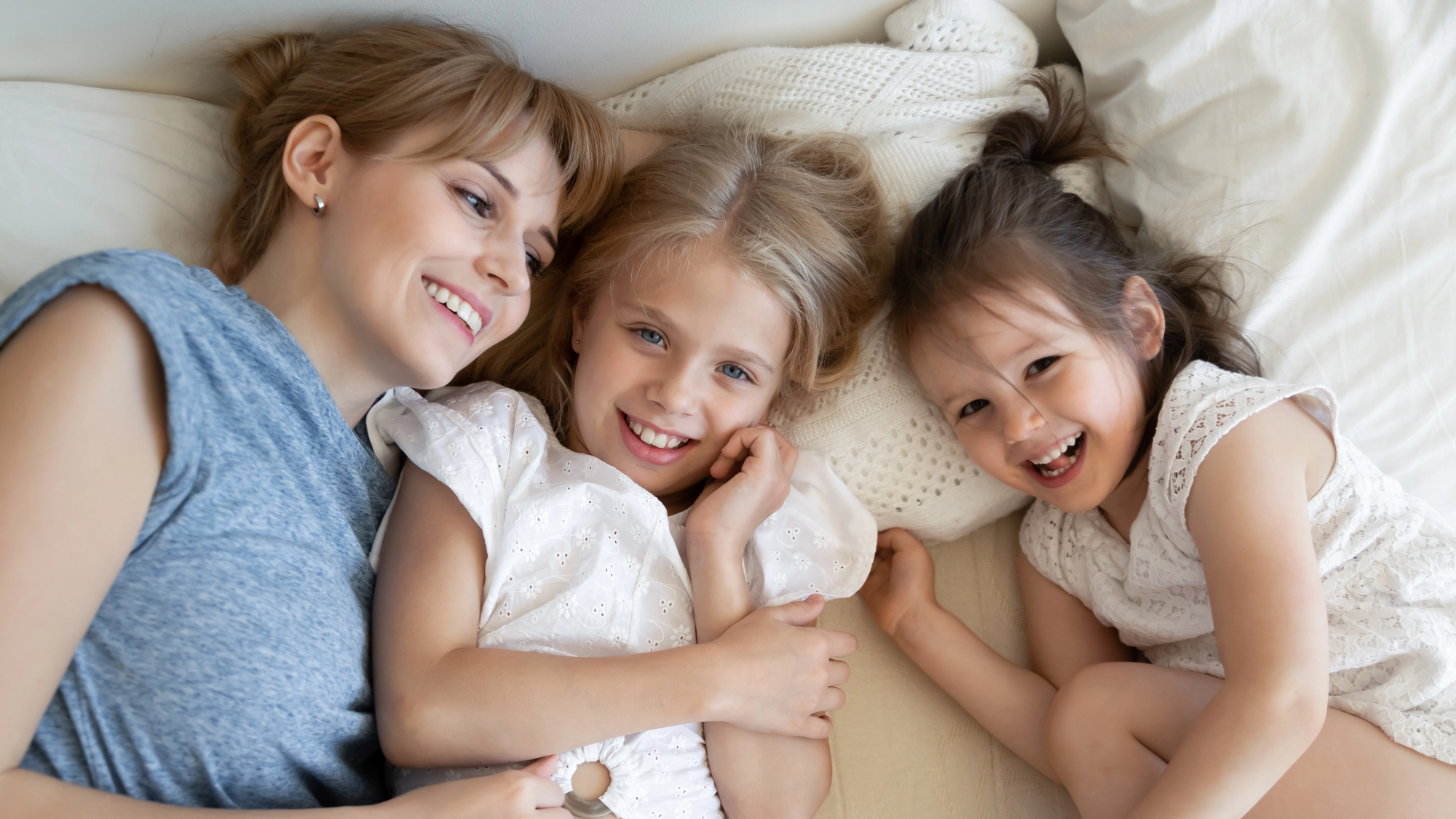 A woman and two young girls are lying on a bed, smiling and laughing. The woman has light brown hair, and the girls have blonde and brown hair respectively. They are dressed in casual, light-colored clothing and appear to be enjoying a joyful moment together.