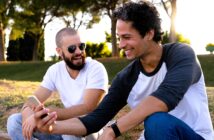 Two men sitting outdoors on a sunny day, smiling and looking at a smartphone together. One man has a beard and wears sunglasses and a white shirt, while the other has short curly hair and wears a baseball-style shirt. Trees are visible in the background.