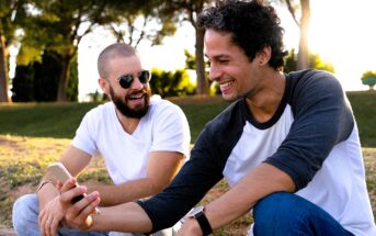 Two men sitting outdoors on a sunny day, smiling and looking at a smartphone together. One man has a beard and wears sunglasses and a white shirt, while the other has short curly hair and wears a baseball-style shirt. Trees are visible in the background.