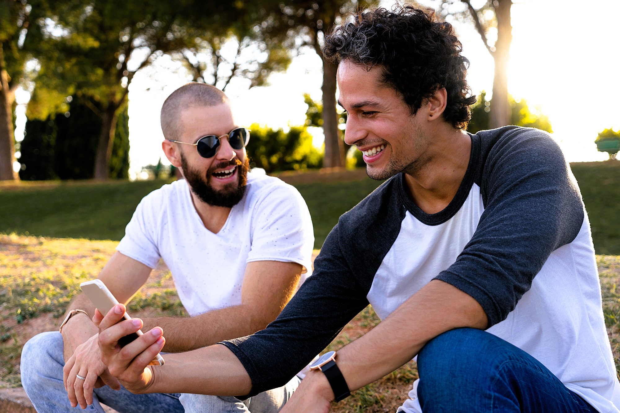 Two men sitting outdoors on a sunny day, smiling and looking at a smartphone together. One man has a beard and wears sunglasses and a white shirt, while the other has short curly hair and wears a baseball-style shirt. Trees are visible in the background.