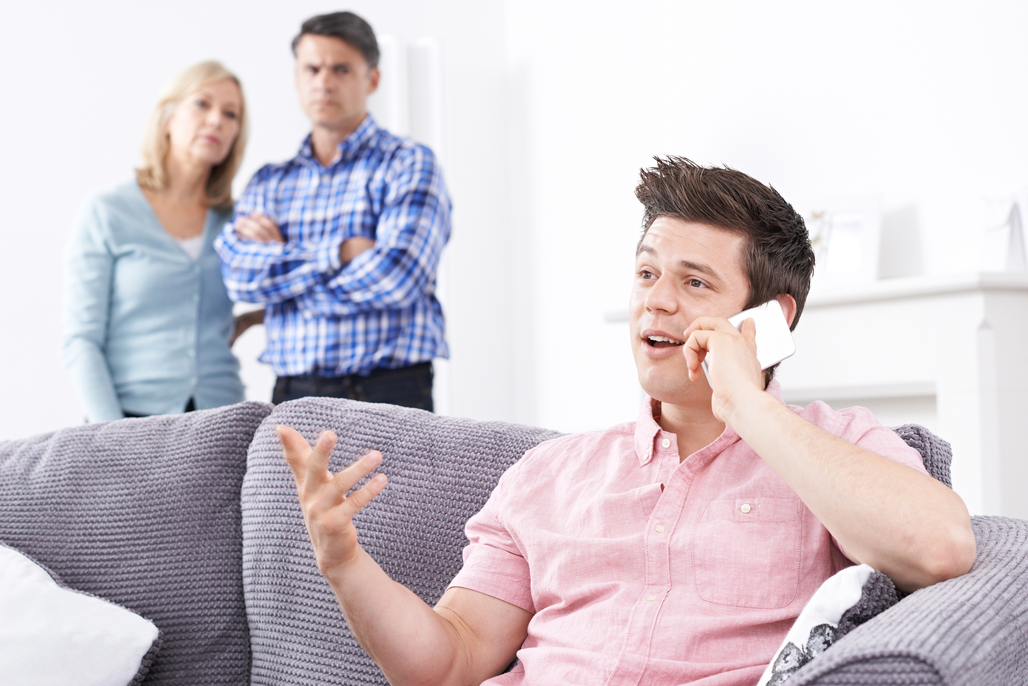 A young man sits on a sofa, talking on his phone, gesturing with his hand. In the background, a middle-aged couple stands together, watching him with arms crossed. The scene suggests a serious or tense conversation.