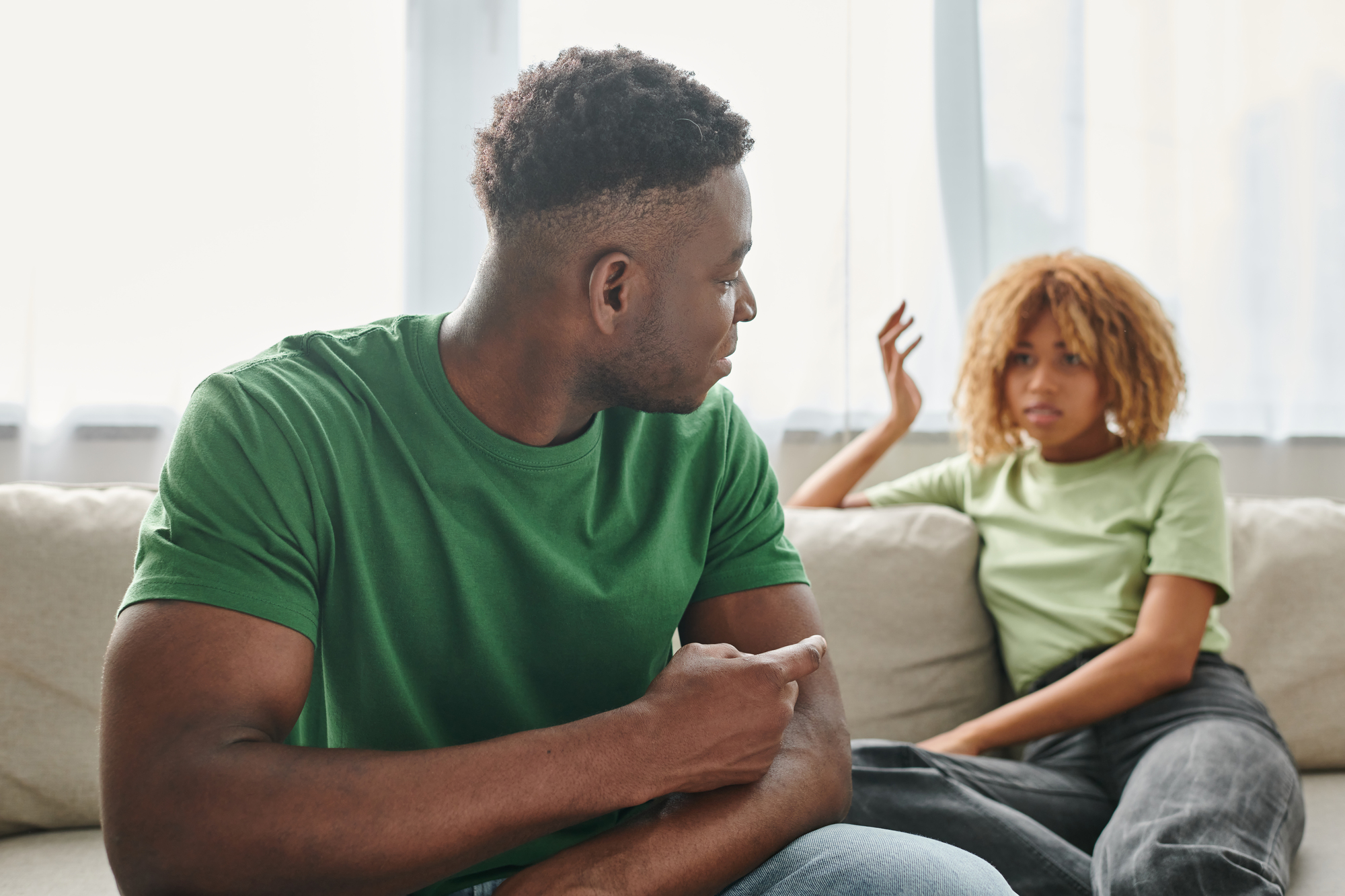 A man and a woman sit on a couch, engaged in a conversation. The man in a green shirt gestures with his hand, while the woman in a light green shirt raises one hand, with a questioning expression. They are in a bright room with large windows.