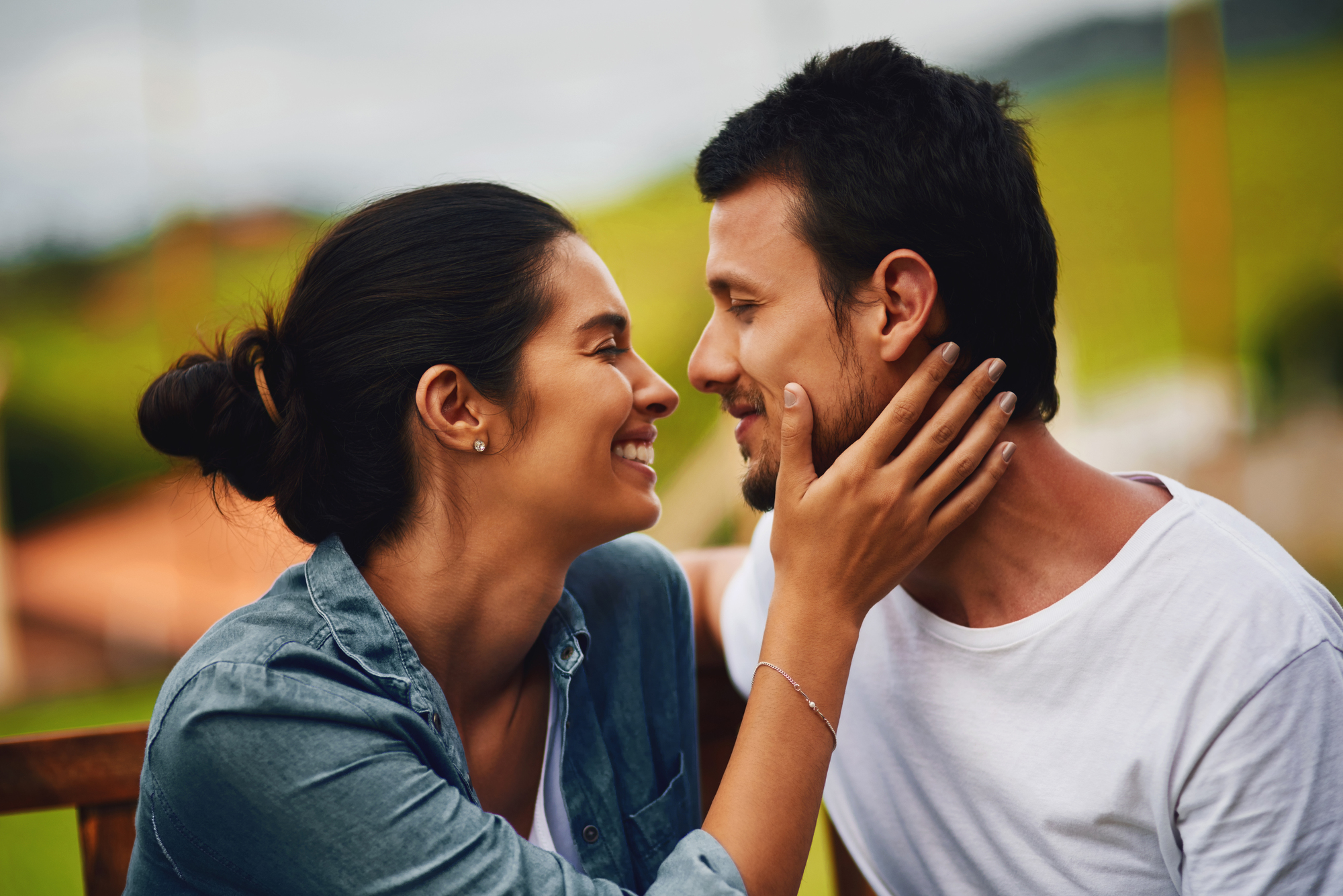 A couple sits closely, smiling at each other lovingly. The woman gently touches the man's face. They are outdoors, with blurred greenery in the background, both appearing happy and relaxed.