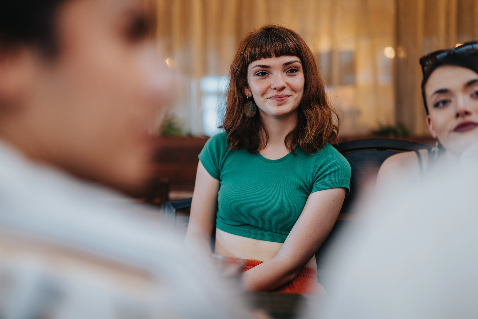 A young woman with shoulder-length brown hair and a green crop top smiles while sitting indoors. She is surrounded by blurry figures, creating a warm and social atmosphere.