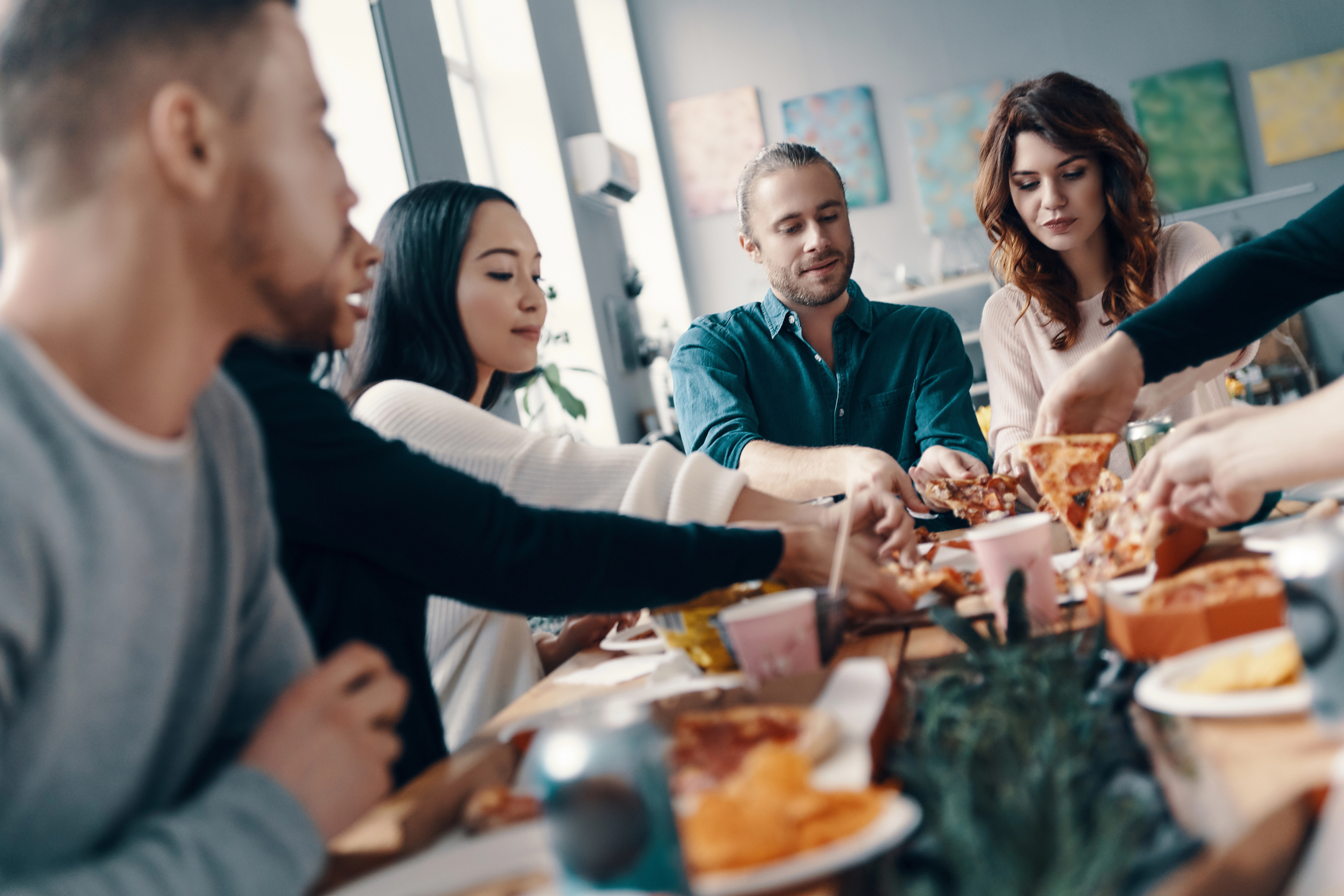 A group of people sitting around a table, enjoying a meal together. They are reaching for various dishes, including pizza and snacks. The setting is casual, with natural light coming in through large windows in the background.