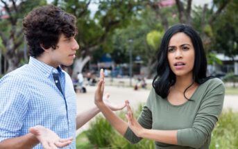 A man and woman are engaged in a heated conversation outdoors in a park. The woman appears frustrated, holding up her hands in a stopping gesture, while the man looks surprised, with his hands open in an expressive manner. Trees are in the background.