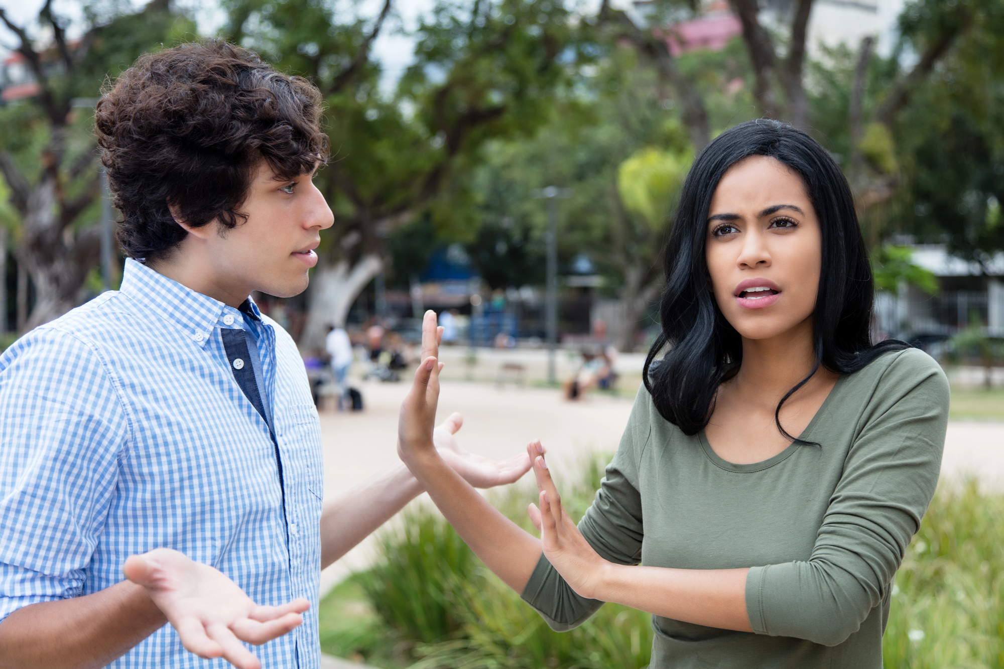 A man and woman are engaged in a heated conversation outdoors in a park. The woman appears frustrated, holding up her hands in a stopping gesture, while the man looks surprised, with his hands open in an expressive manner. Trees are in the background.