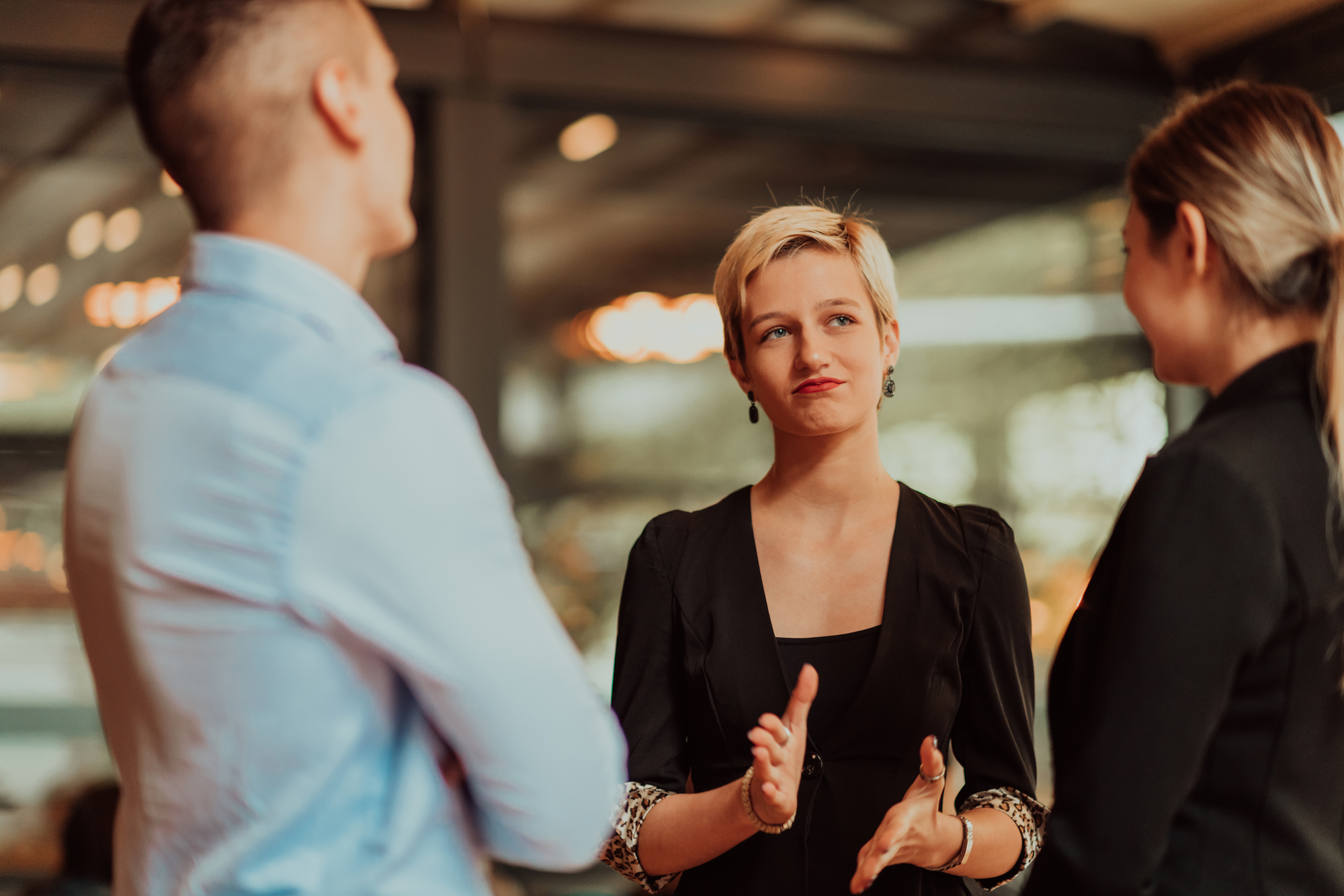 A group of three people engaged in conversation indoors. The person in the middle is gesturing with hands, wearing a black outfit. The background is softly blurred, suggesting a casual or business setting.