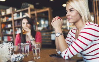 Two women sitting at a wooden table in a cafe, chatting. The woman in the foreground wears a striped top and a watch, while the one behind drinks from a glass. The setting is cozy, with shelves and warm lighting in the background.