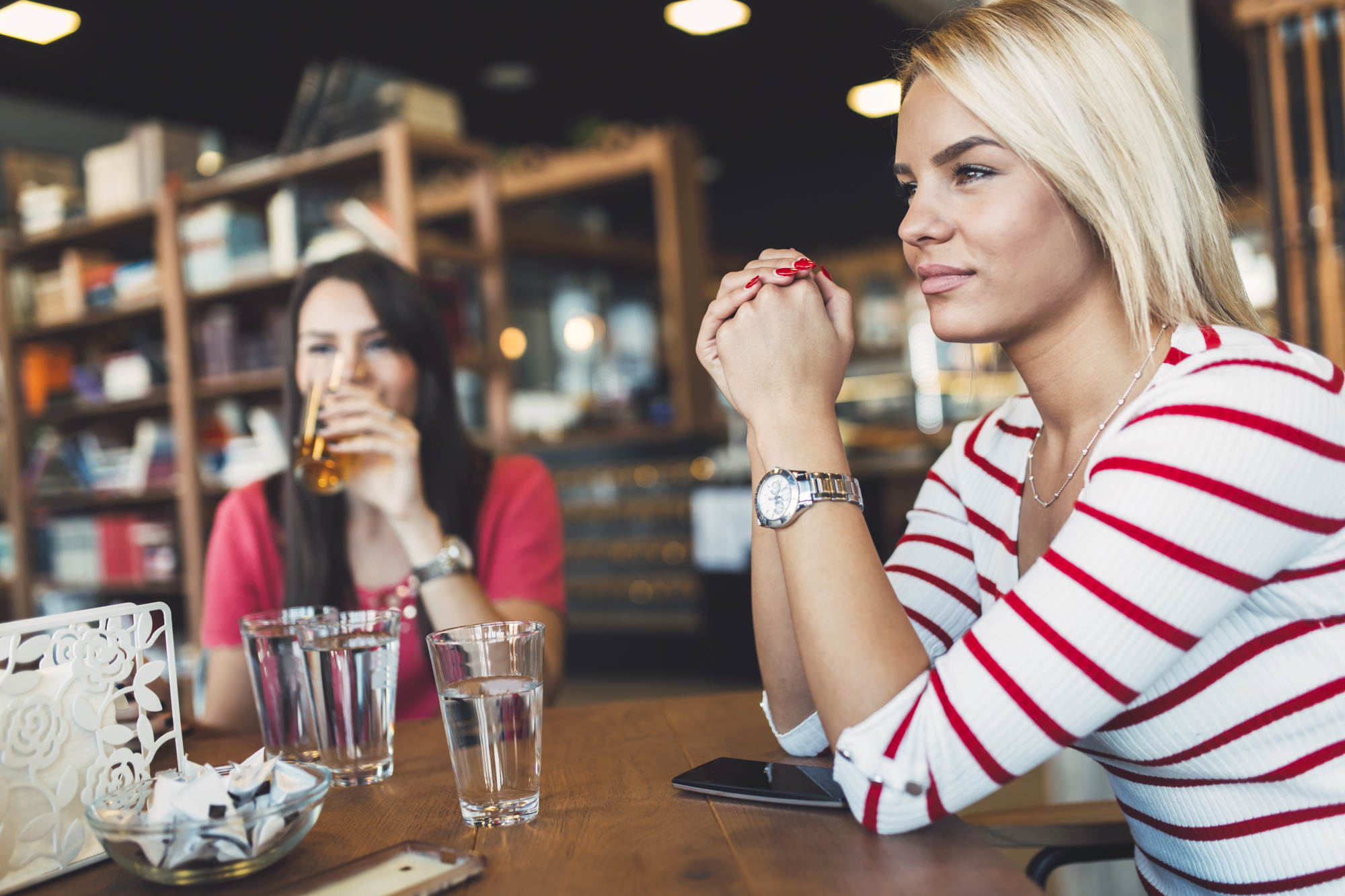 Two women sitting at a wooden table in a cafe, chatting. The woman in the foreground wears a striped top and a watch, while the one behind drinks from a glass. The setting is cozy, with shelves and warm lighting in the background.