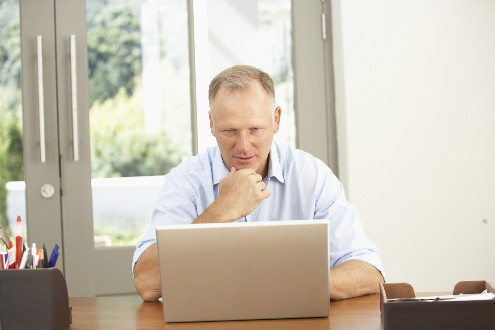 A man sits at a desk looking at a laptop. He is wearing a light blue shirt and has his hand on his chin in thought. The background shows a window with a view of greenery outside. There are pens and office supplies on the desk.