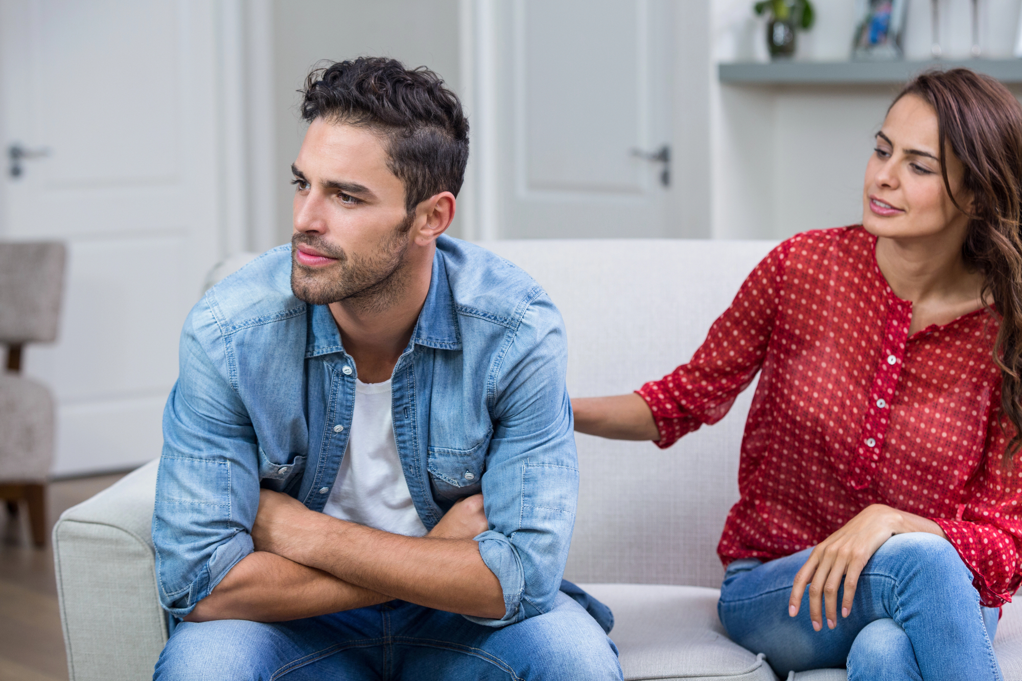 A man sitting on a couch with arms crossed, looking away, while a woman in a red blouse sits beside him, gently touching his shoulder. The setting appears to be a bright living room, suggesting a serious or emotional conversation.